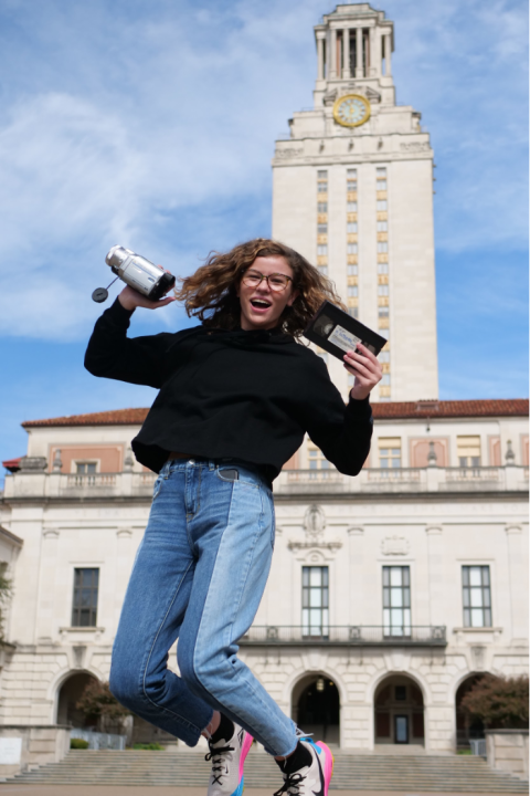 Photo is of a young girl jumping up in the air while holding a video camera in her right hand and a tape in her left hand. In the background shows the UT Austin Tower behind her. 