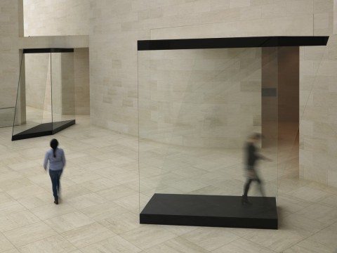 Two glass and wood structures in the atrium of a museum. A person can be seen walking through one and another person is walking towards another. Both figures are slightly blurred showing movement. 
