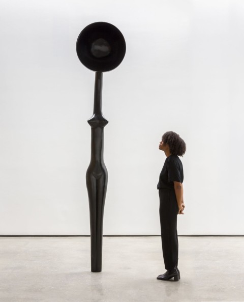 An image of Simone Leigh's "Sentinel IV" in a blank white gallery space with an onlooking black female figure. The work is cast in dark bronze and resembles an elongated female form with a large bowl shape as a head.