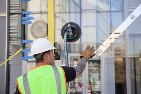 A construction worker wearing a hard hat and vest guides a large, semi-reflective piece of glass suspended in the air 