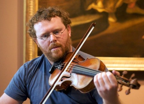 An Image of Travis Weller playing a violin. He wears glasses and a blue shirt.