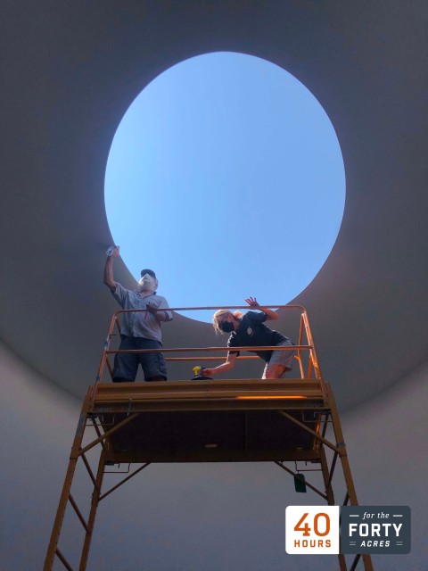 Two people stand on a scaffold in James Turrell's skyspace "The Color Inside" and they stand against the blue sky framed by the oculus.