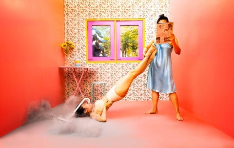 A still from Kawita Vatanajyankur's "Tools (video series); Two figures are seen in this still. One person reading a piece of paper supports another person like a vacuum. The room is pink with wallpaper in the center with a window.