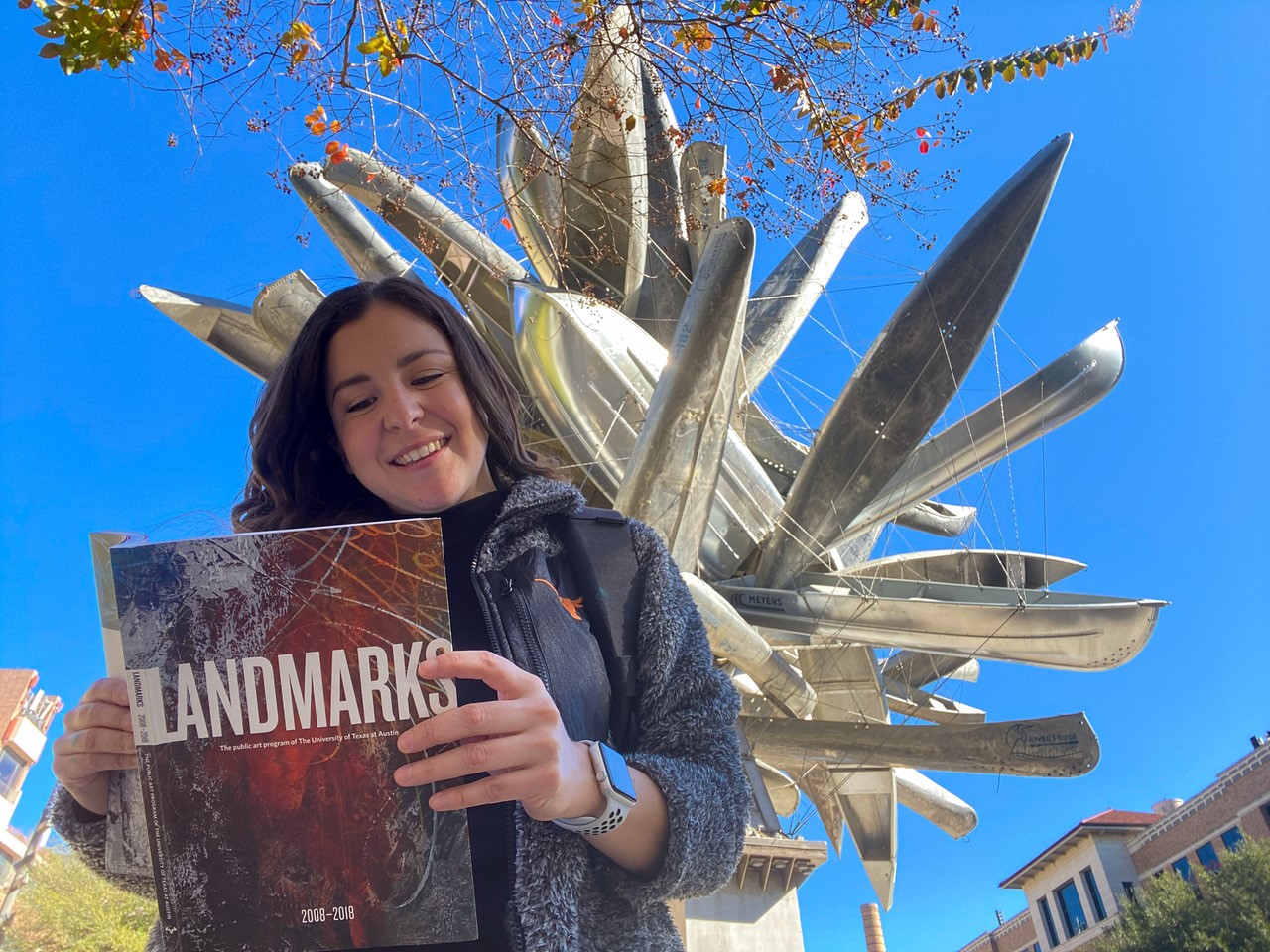 A woman with dark brown hair looks down at a Landmarks Handbook. She wears a UT jacket with a "longhorn" emblem on the front. She stands in front of Nancy Rubins' "Monochrome for Austin" a sculpture made of canoes assembled and suspended in air.