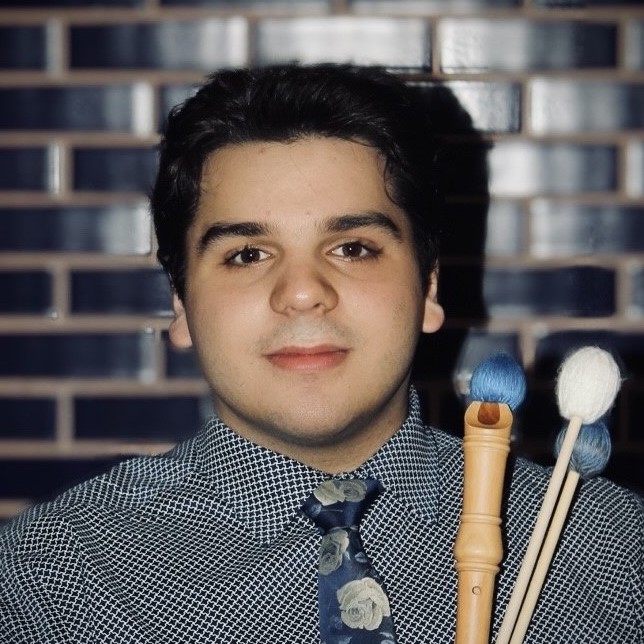 A photograph of Josiah Garza; He looks at the camera, wears a floral tie, and holds musical instruments